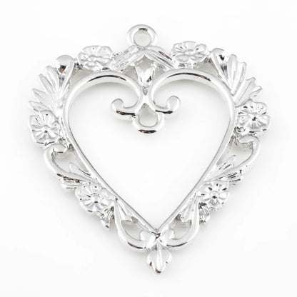 Silver heart-shaped jewelry pendant with an ornate filigree border, floral details, and a top loop, shown on a plain white background.