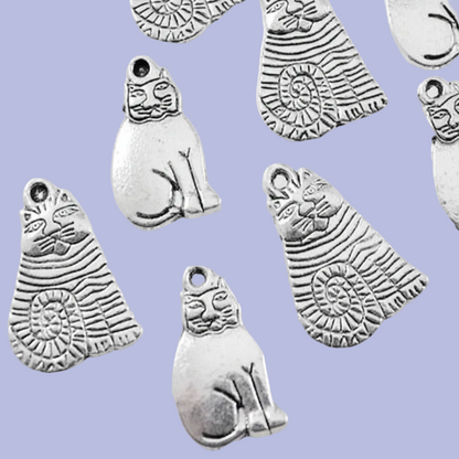 Multiple silver-toned metal charms shaped like stylized cats with carved facial features and striped body patterns, scattered on a light lavender background.