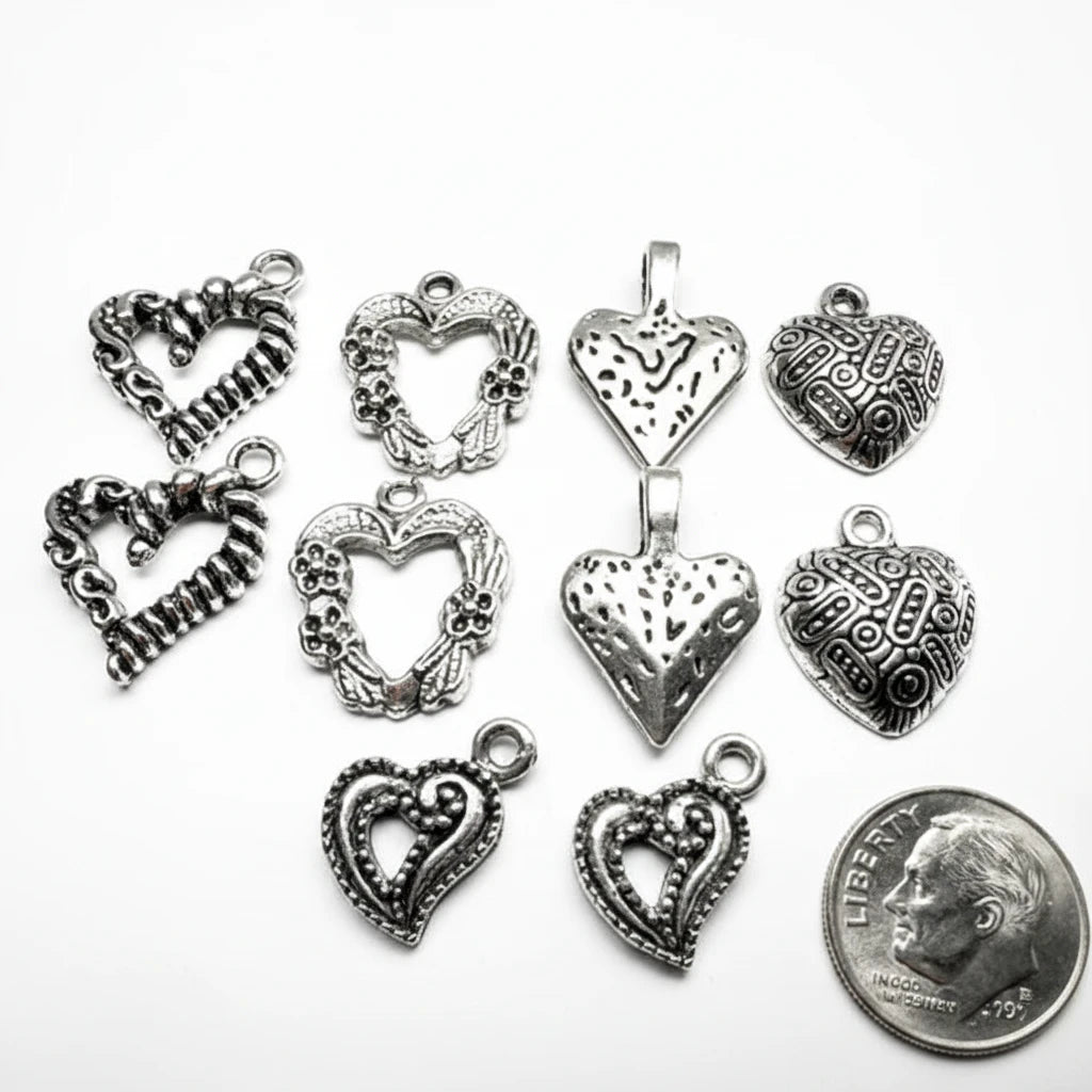 Nine silver-toned heart-shaped jewelry charms with various textured, filigree, and open heart designs are arranged on a white background next to a US quarter coin for scale.
