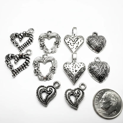 Nine silver-toned heart-shaped jewelry charms with various textured, filigree, and open heart designs are arranged on a white background next to a US quarter coin for scale.