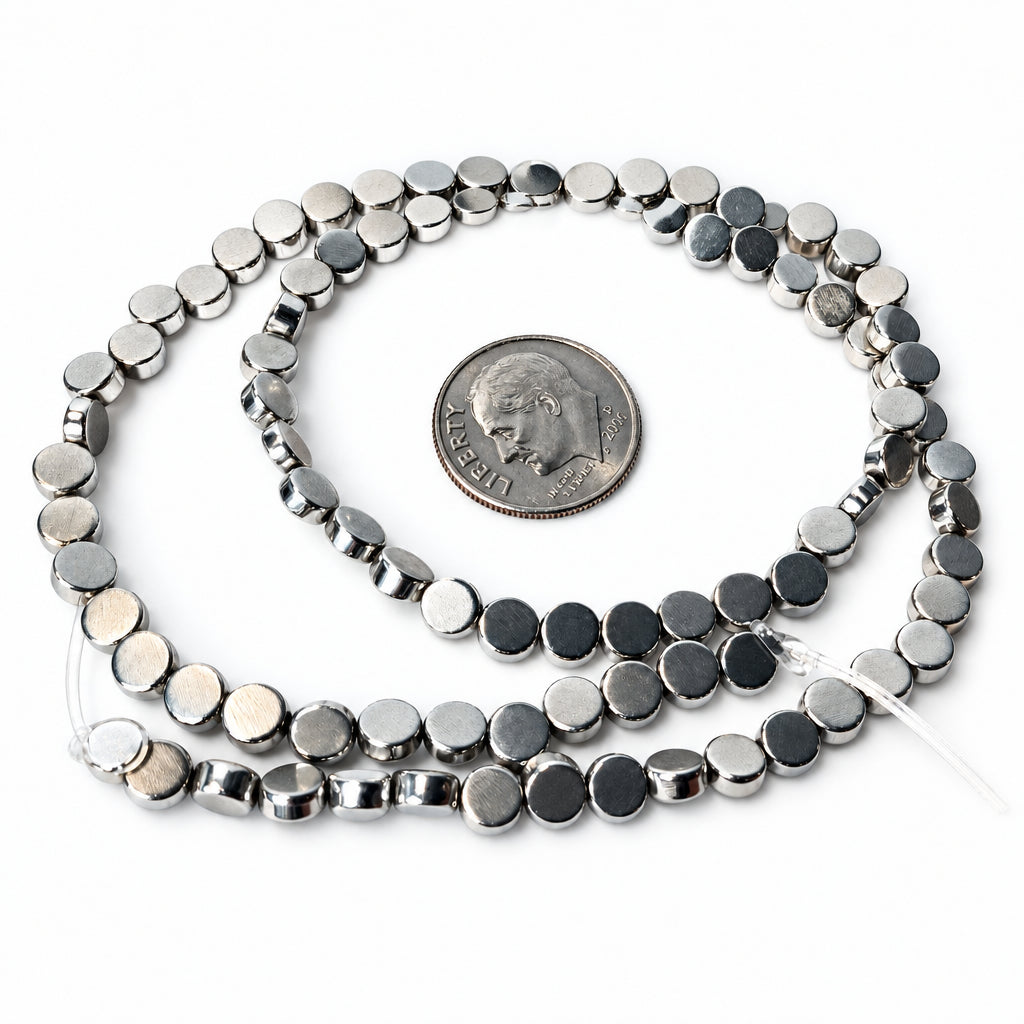 A strand of small metallic silver disc beads coiled on a white surface, with a U.S. dime placed in the center for scale.