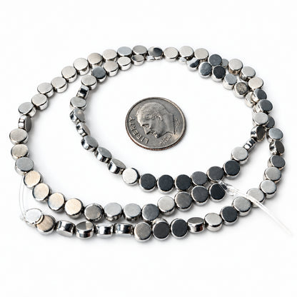 A strand of small metallic silver disc beads coiled on a white surface, with a U.S. dime placed in the center for scale.