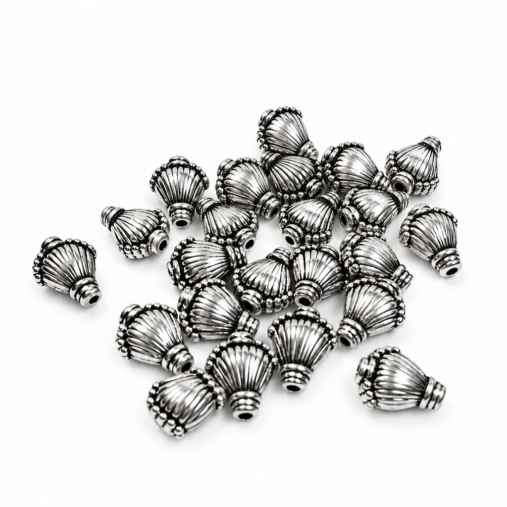 A pile of small metallic silver decorative beads with a textured, shell  ribbed design, scattered on a plain white background.