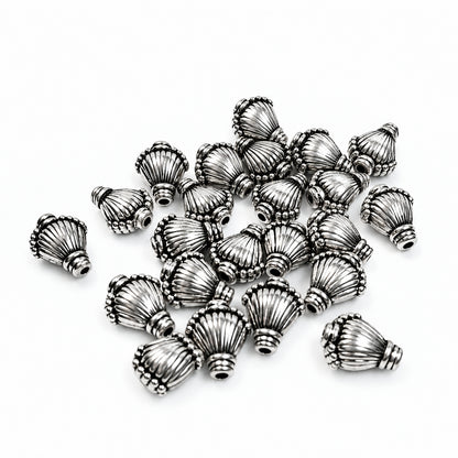 A pile of small metallic silver decorative beads with a textured, shell  ribbed design, scattered on a plain white background.