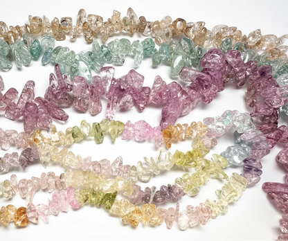 A collection of multicolor raw crystal chip beads arranged in overlapping rows on a white background, showing shades of pink, purple, green, yellow, and orange.