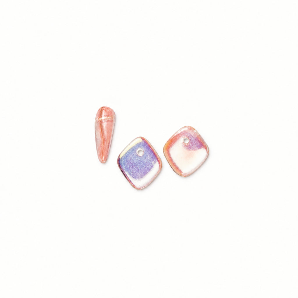 Three iridescent resin beads shown on a plain white background: one elongated teardrop bead, one square bead with a lavender-purple center, and one square bead with a pink-peach center.