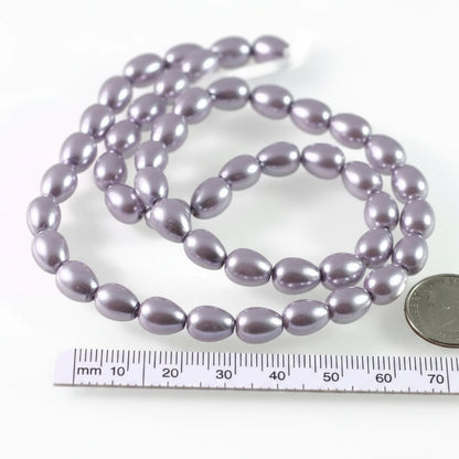 A strand of glossy, teardrop-shaped beads in a dusty lavender-purple color, displayed on a white background with a ruler and a coin for scale.