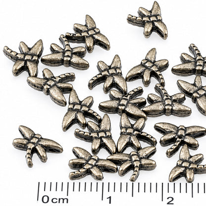 A group of small metallic dragonfly shaped craft charms or beads in an antique bronze finish, arranged on a plain white background with a 0-3 cm ruler at the bottom for scale.