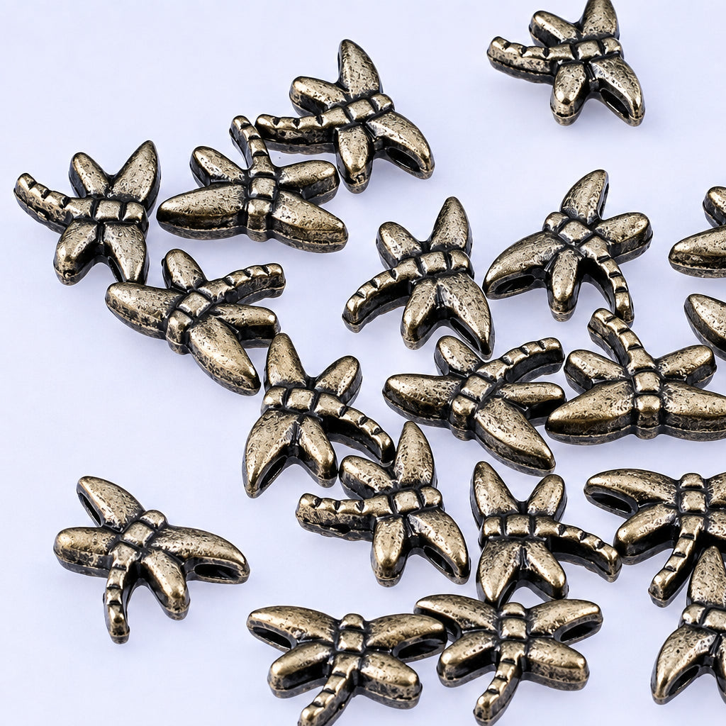A pile of small metallic dragonfly-shaped beads or charms with an antique bronze finish, scattered on a plain light background.