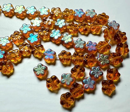 A strand of translucent amber glass beads shaped like four-leaf clovers, with some beads featuring iridescent pastel blue and white centers, displayed on a plain white surface.