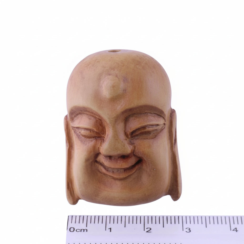 A carved wooden Buddha head pendant or ornament with a smooth, bald head and closed eyes, shown against a light purple background with a 0-3 cm ruler at the bottom for scale.
