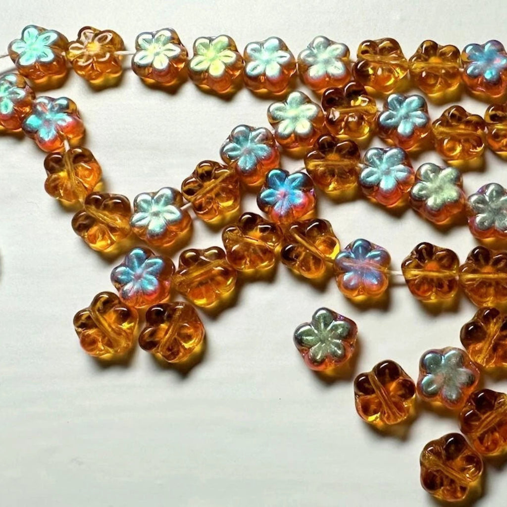 A strand of small flower-shaped beads in amber orange with iridescent pastel accents, laid out on a plain white surface.