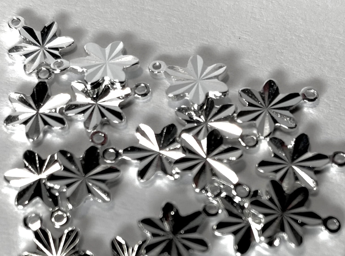 A pile of silver-toned metal flower-shaped charms with small loops at the top, photographed on a light gray textured background.