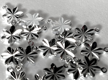 A pile of silver-toned metal flower-shaped charms with small loops at the top, photographed on a light gray textured background.