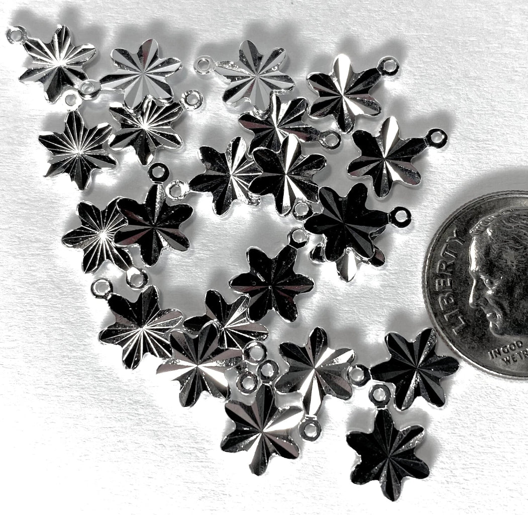 A cluster of silver-toned metal snowflake charms with small loops at the top, arranged on a white textured surface next to a US quarter for scale.