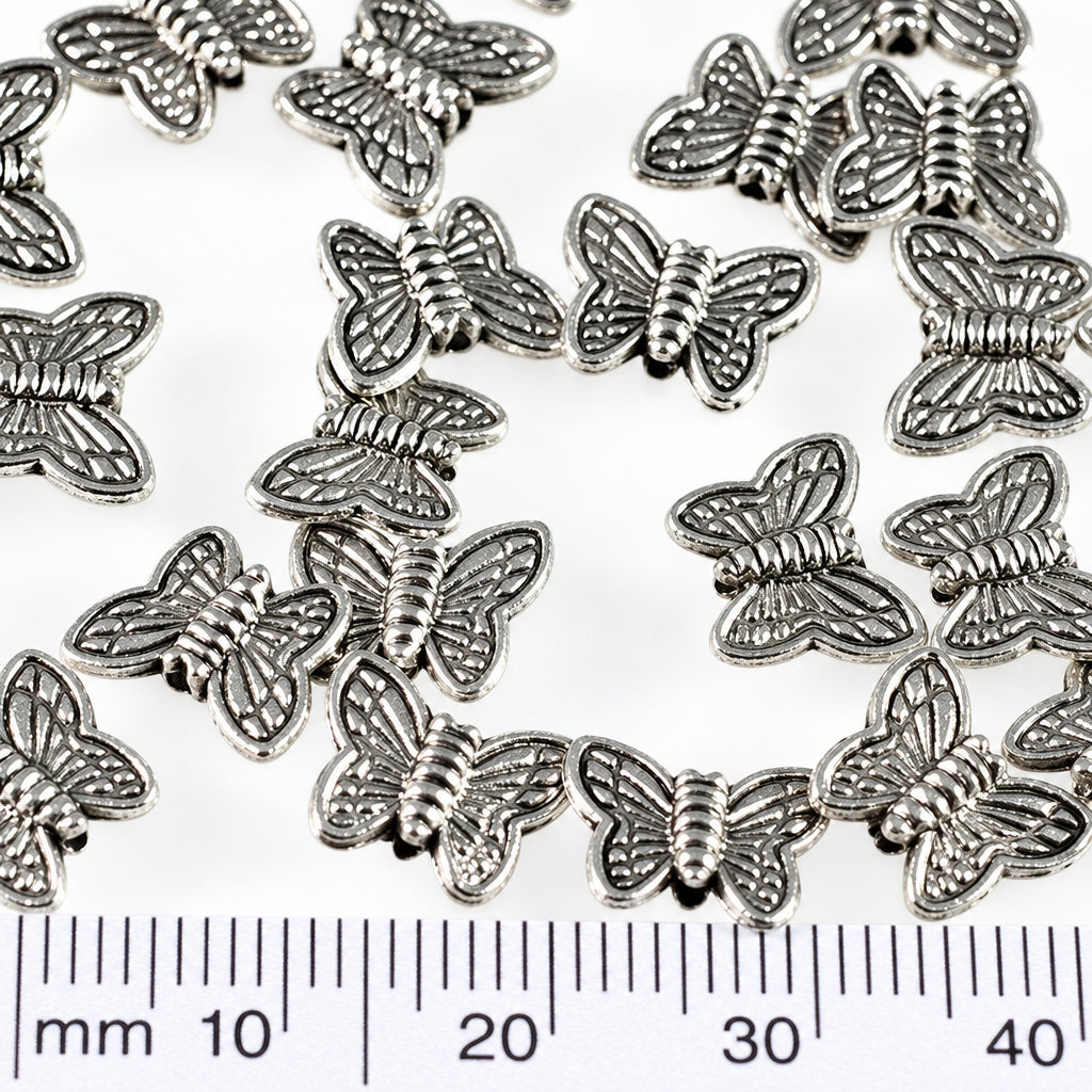 Multiple small silver butterfly-shaped findings scattered on a white background, with a ruler at the bottom showing millimeter measurements and a coin at the right for scale.
