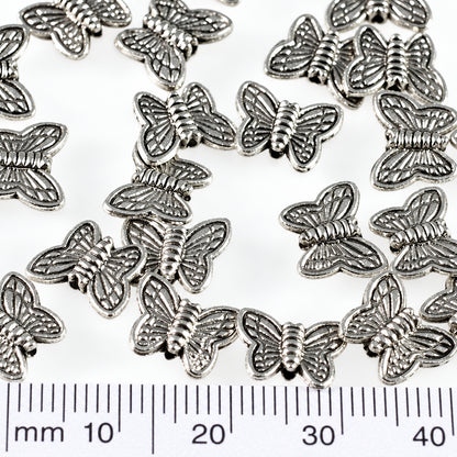 Multiple small silver butterfly-shaped findings scattered on a white background, with a ruler at the bottom showing millimeter measurements and a coin at the right for scale.