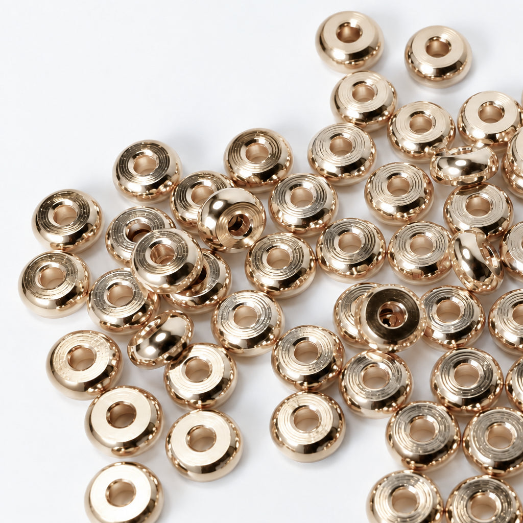 A pile of small round metallic gold spacers or beads with a central hole, scattered on a plain white surface.