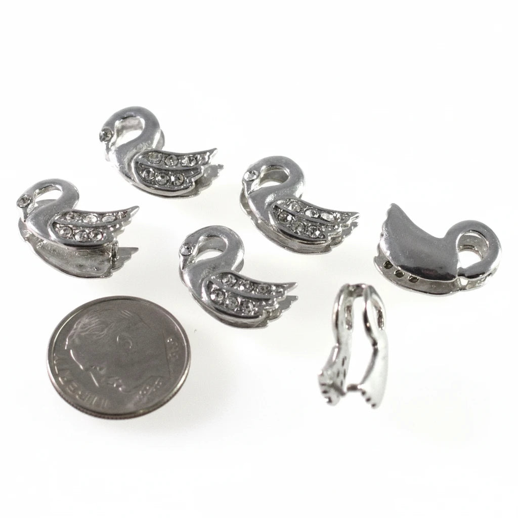 Five silver-toned swan-shaped jewelry findings or charms, some showing clear rhinestone detailing, displayed on a white background with a US quarter coin for scale.