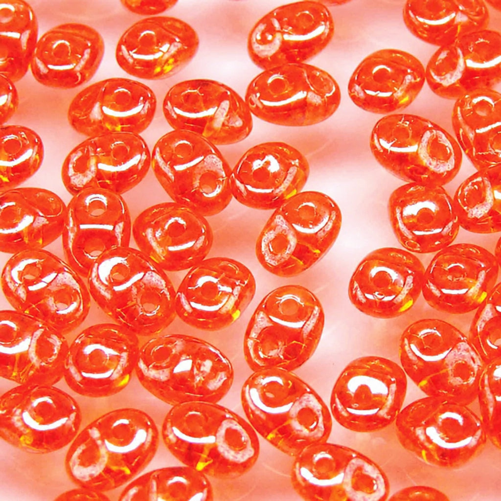 A close-up of many small translucent orange-red oval beads scattered across a pale pink background, with a white outline drawing of a test tube overlaid in the center.