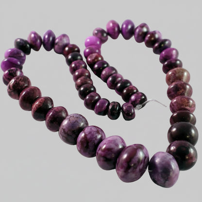A strand of polished purple and black marbled beads, likely chrysocolla, arranged in a loop on a white textured background.