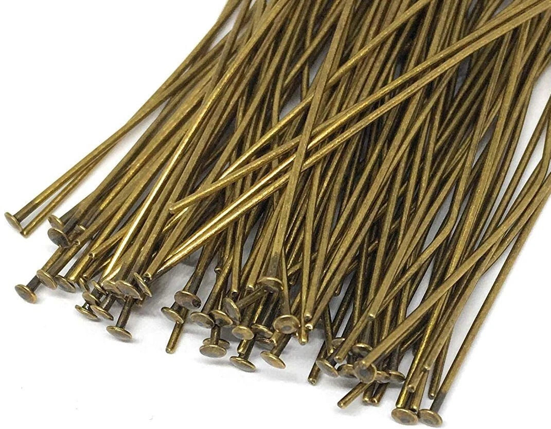 A pile of long, thin metallic pins with round heads, finished in a warm antique gold or bronze tone, arranged on a plain white background.