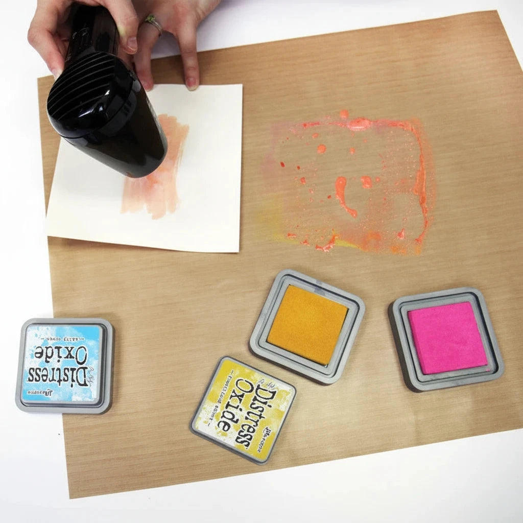 A top-down view of a person using a black hair dryer to dry an orange ink stamp on white paper, placed on a wooden board. Three Distress Oxide ink pads in blue, yellow, and pink are visible in the foreground.