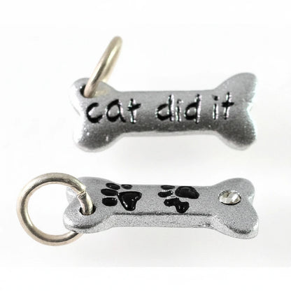 Two silver-tone metal bone-shaped charms on a plain white background. The top charm is engraved with the words 'cat did it' and has a small hanging loop at the top.