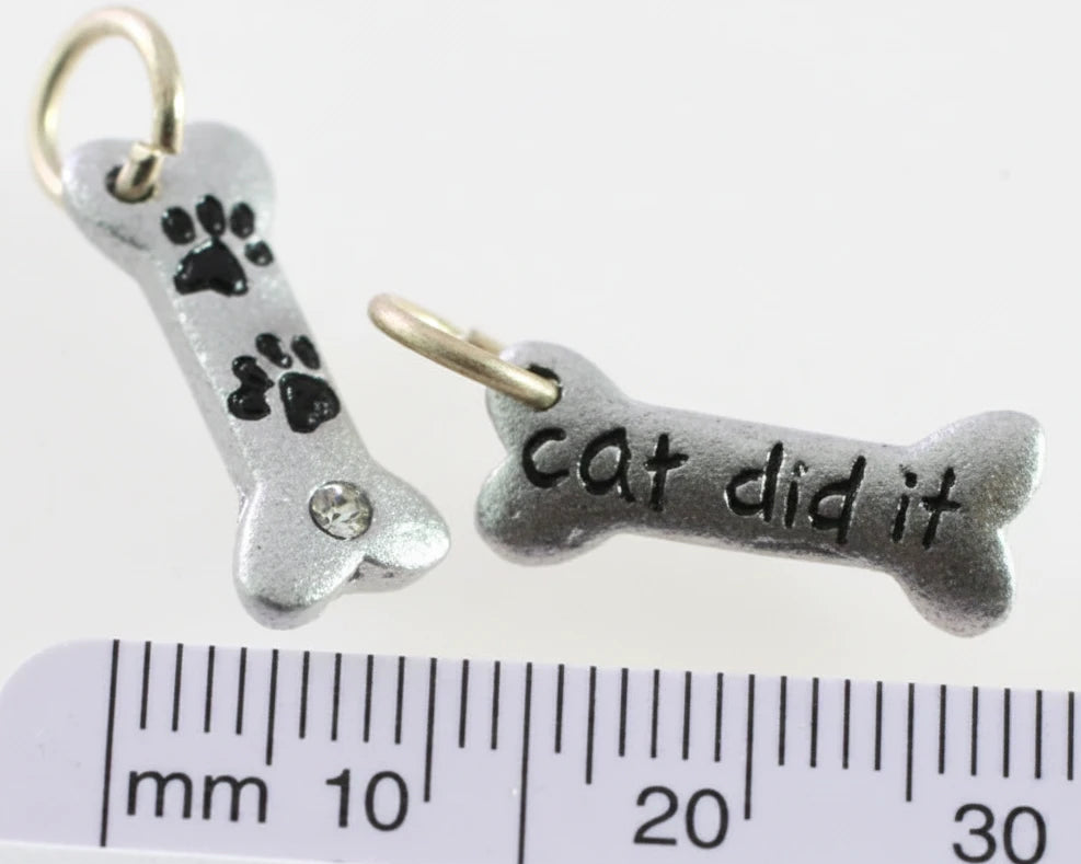 Two silver-toned bone-shaped charms with gold-tone loops, shown above a ruler for scale. One charm has black paw prints and a small clear rhinestone, and the other reads 'cat did it' in black lettering.