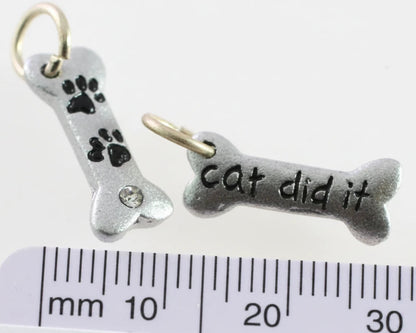 Two silver-toned bone-shaped charms with gold-tone loops, shown above a ruler for scale. One charm has black paw prints and a small clear rhinestone, and the other reads 'cat did it' in black lettering.