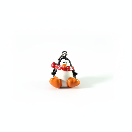 A small penguin charm with a black and white body, orange beak and feet, and a red scarf with white polka dots, attached to a silver jump ring on a white background.