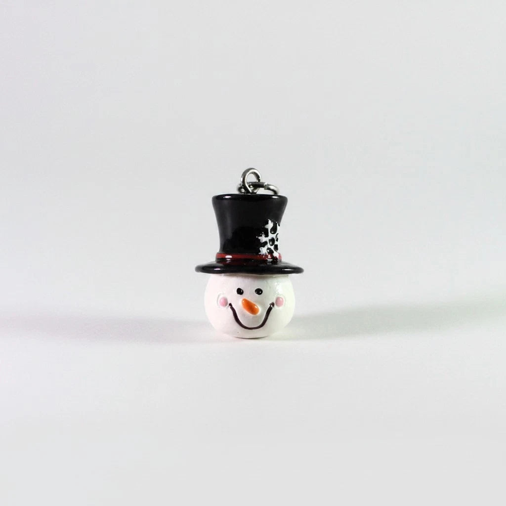 A small snowman ornament or charm with a white round body, black top hat decorated with white polka dots, and a black scarf, shown on a plain light gray background.