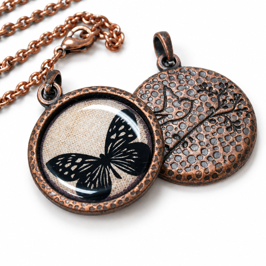 Round copper-toned pendant necklace shown open to reveal both sides: one side features a black butterfly design over a textured beige background, and the other side has a hammered floral pattern. A chain is visible in the upper left on a light wood surface.