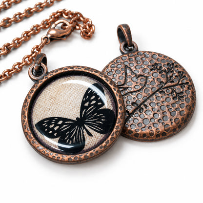 Round copper-toned pendant necklace shown open to reveal both sides: one side features a black butterfly design over a textured beige background, and the other side has a hammered floral pattern. A chain is visible in the upper left on a light wood surface.