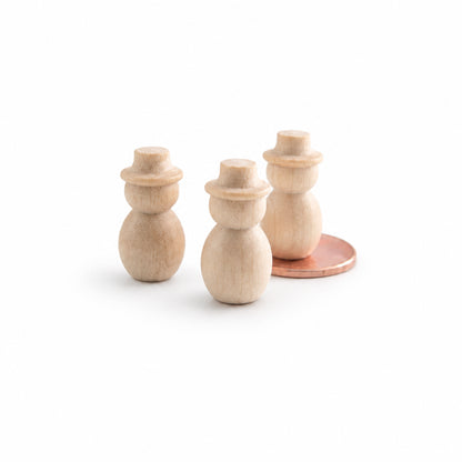 Three small unfinished wooden snowman figurines with a warm natural wood finish, displayed on a clean white reflective surface with a copper coin behind the rightmost figure for scale.