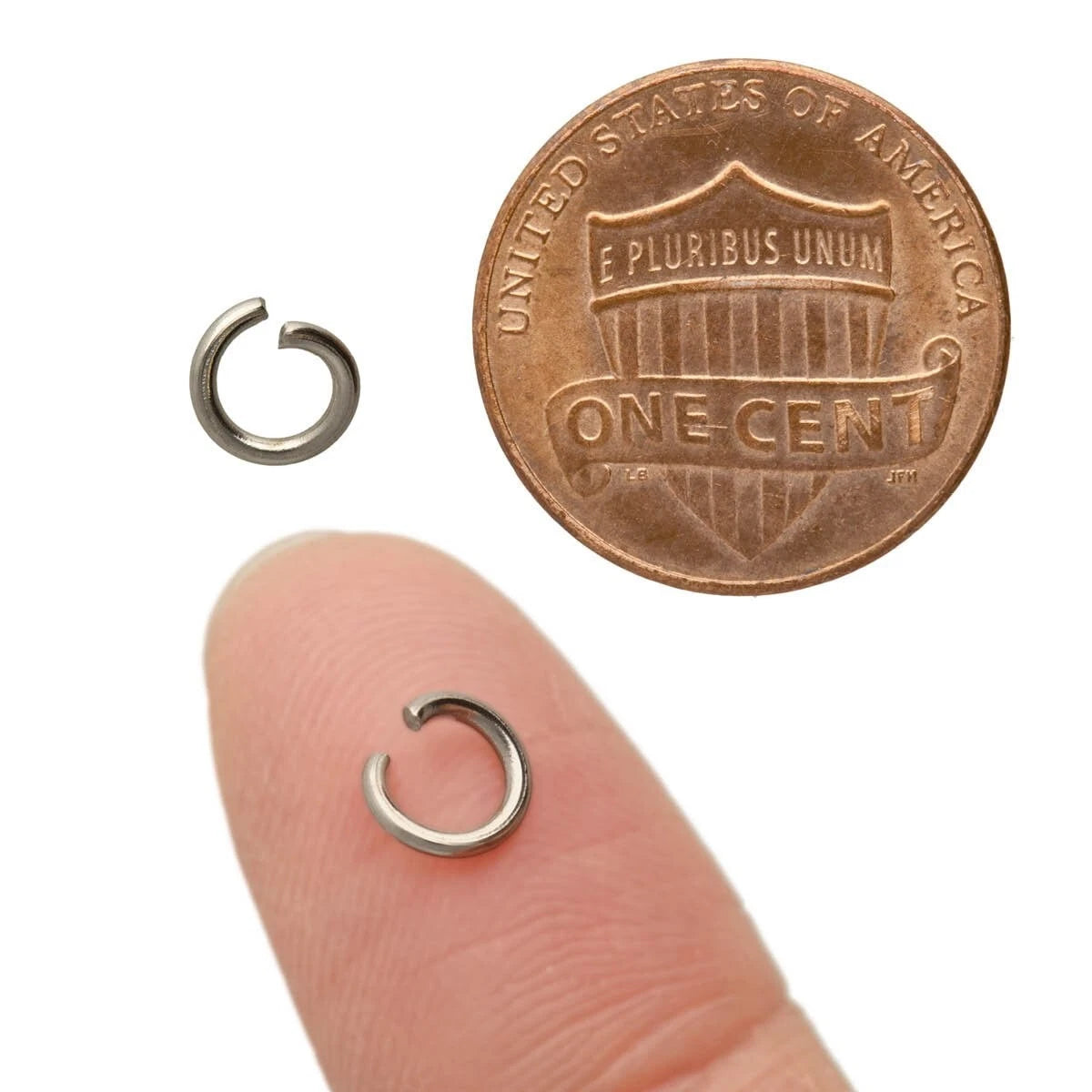 Two small silver metal jump rings shown on a white background, with one near a penny for scale and the other resting on a human fingertip to demonstrate size.