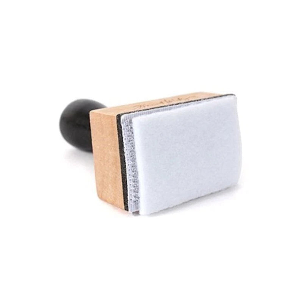 A small rectangular stamp or applicator tool with a black round handle, a light wood block, and a white textured pad, shown on a plain white background.