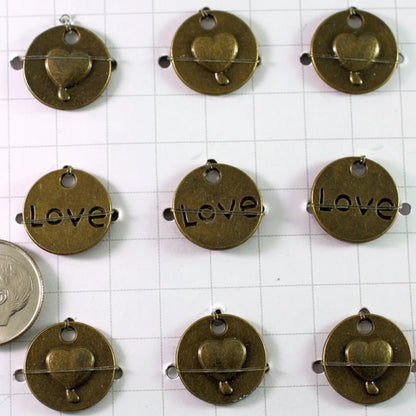Nine round antique bronze metal charms with heart and acorn motifs, three featuring the word 'Love' engraved, arranged on a white grid background next to a coin for scale.