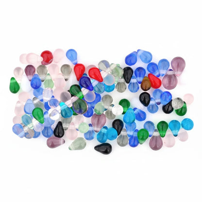 A close-up of many small teardrop-shaped translucent beads in assorted colors including blue, green, red, black, pink, purple, and clear, scattered across a white background.