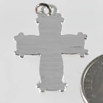 A silver cross pendant with a textured finish and a small hanging loop at the top, shown on a light gray background with a coin partially visible at the bottom right for scale.