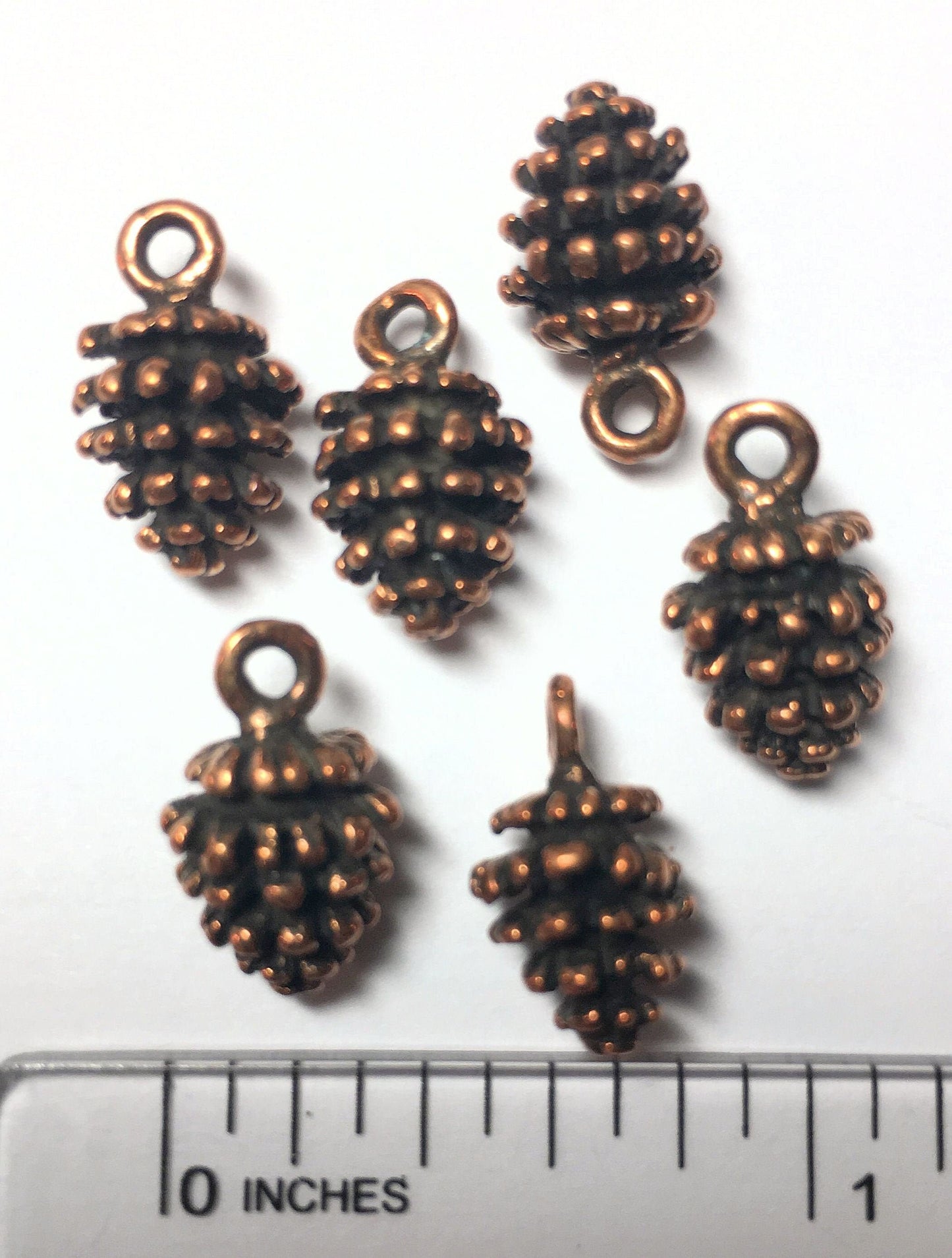 Set of copper-colored pine cone charms with a ruler for scale on a white background