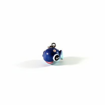 A small blue fish-shaped charm or pendant with a shiny finish, featuring large cartoon eyes, a red mouth, beige spots, and a silver metal loop at the top, shown on a plain white background.
