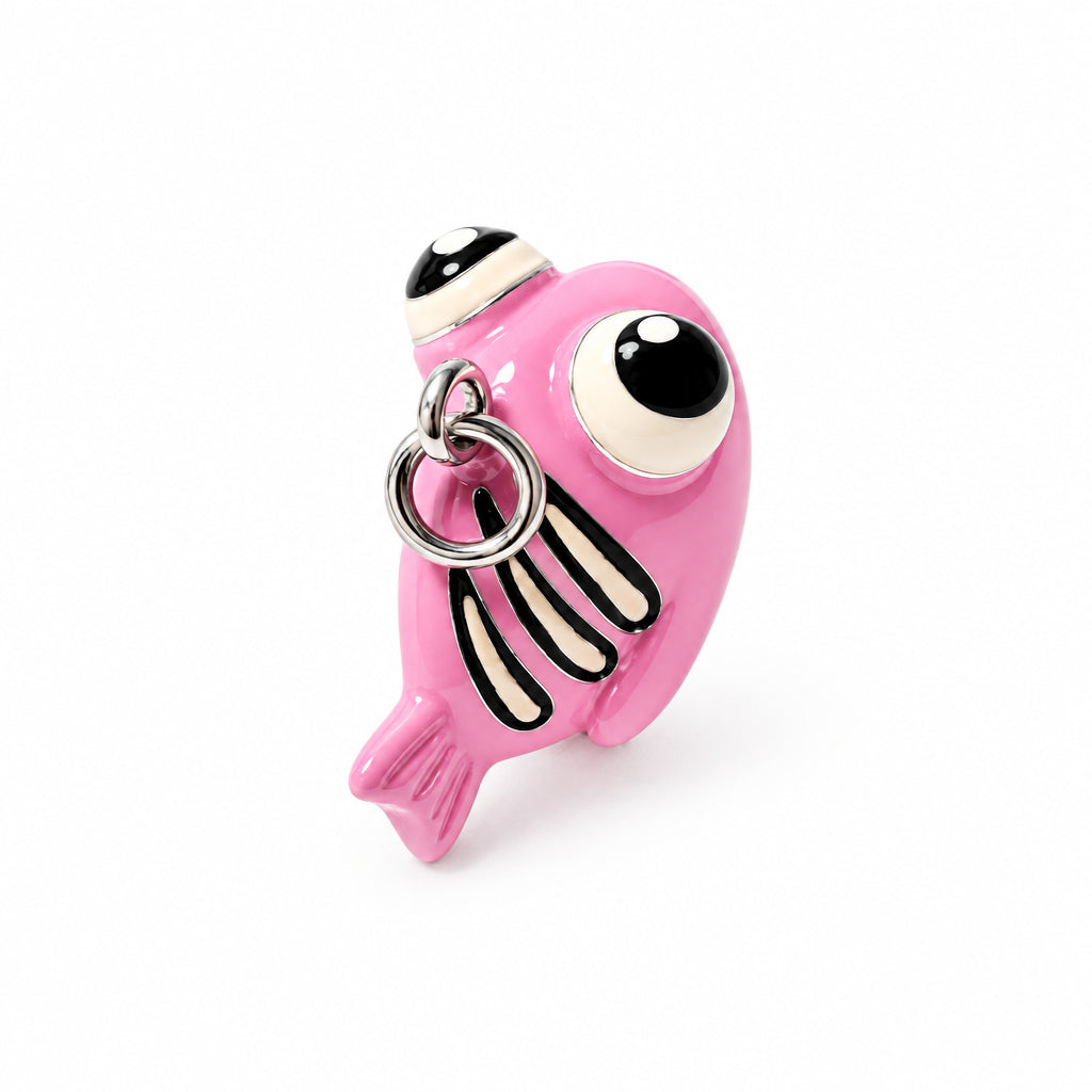 A pink fish-shaped charm or keychain with a large black-and-white eye, black vertical stripes, and a silver metal ring, shown on a plain light background.