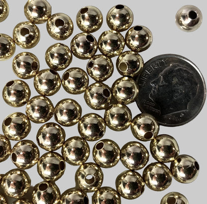 A pile of small metallic gold round beads with central holes, shown on a white surface next to a U.S. dime for scale.