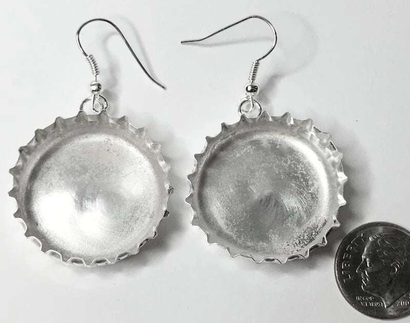 A pair of handmade silver bottle cap earrings with fishhook wires, displayed on a white background next to a US quarter coin for scale.