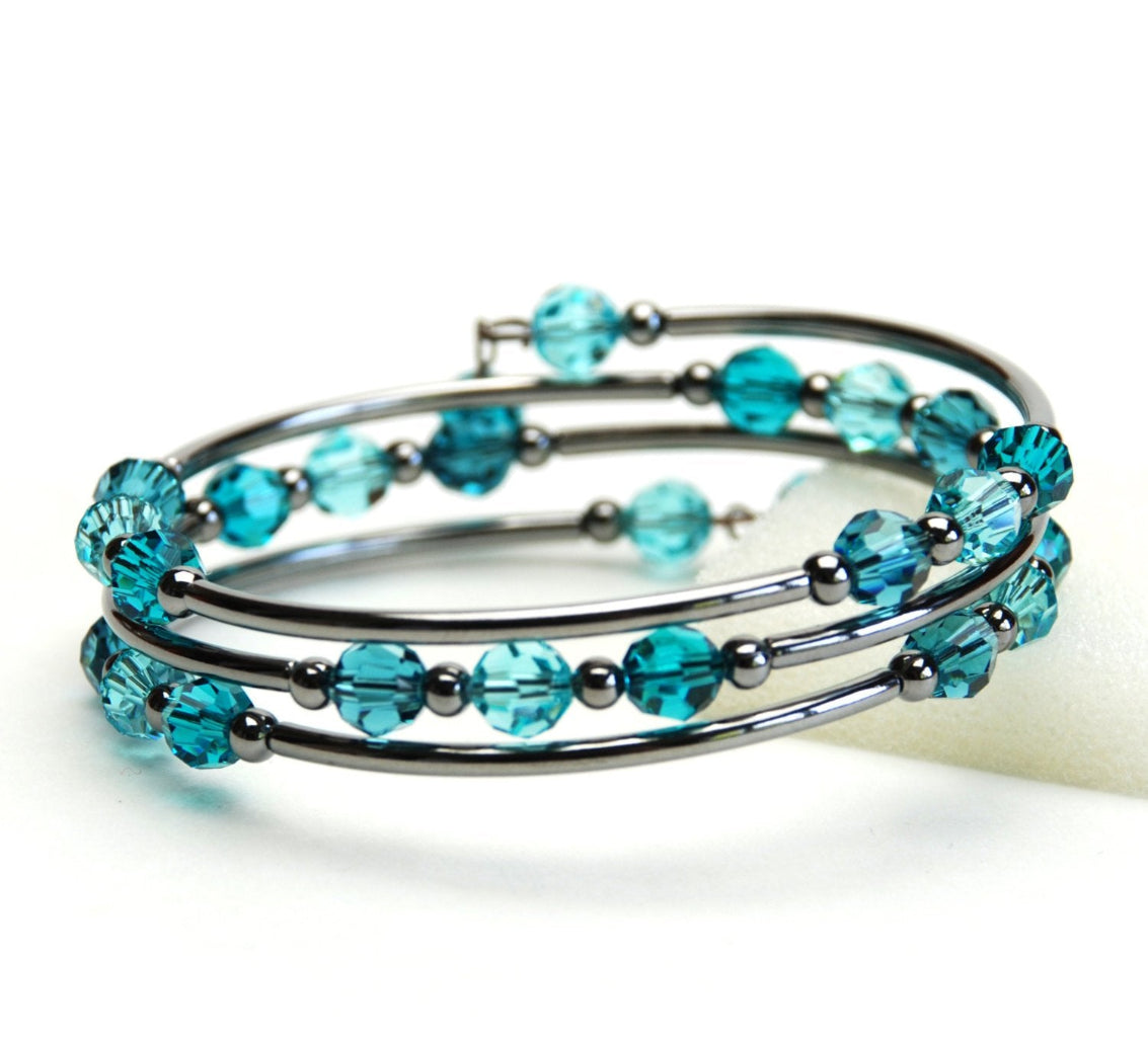 A close-up studio shot of a multi-strand silver-toned bangle bracelet adorned with faceted teal-blue glass beads, resting on a white surface.