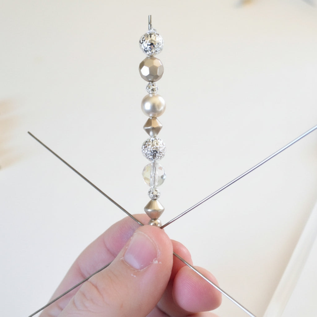 A close-up of a hand holding a beaded wire pick with metallic gold and silver beads, with a pink hot air gun and loose beads in the background.