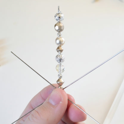 A close-up of a hand holding a beaded wire pick with metallic gold and silver beads, with a pink hot air gun and loose beads in the background.